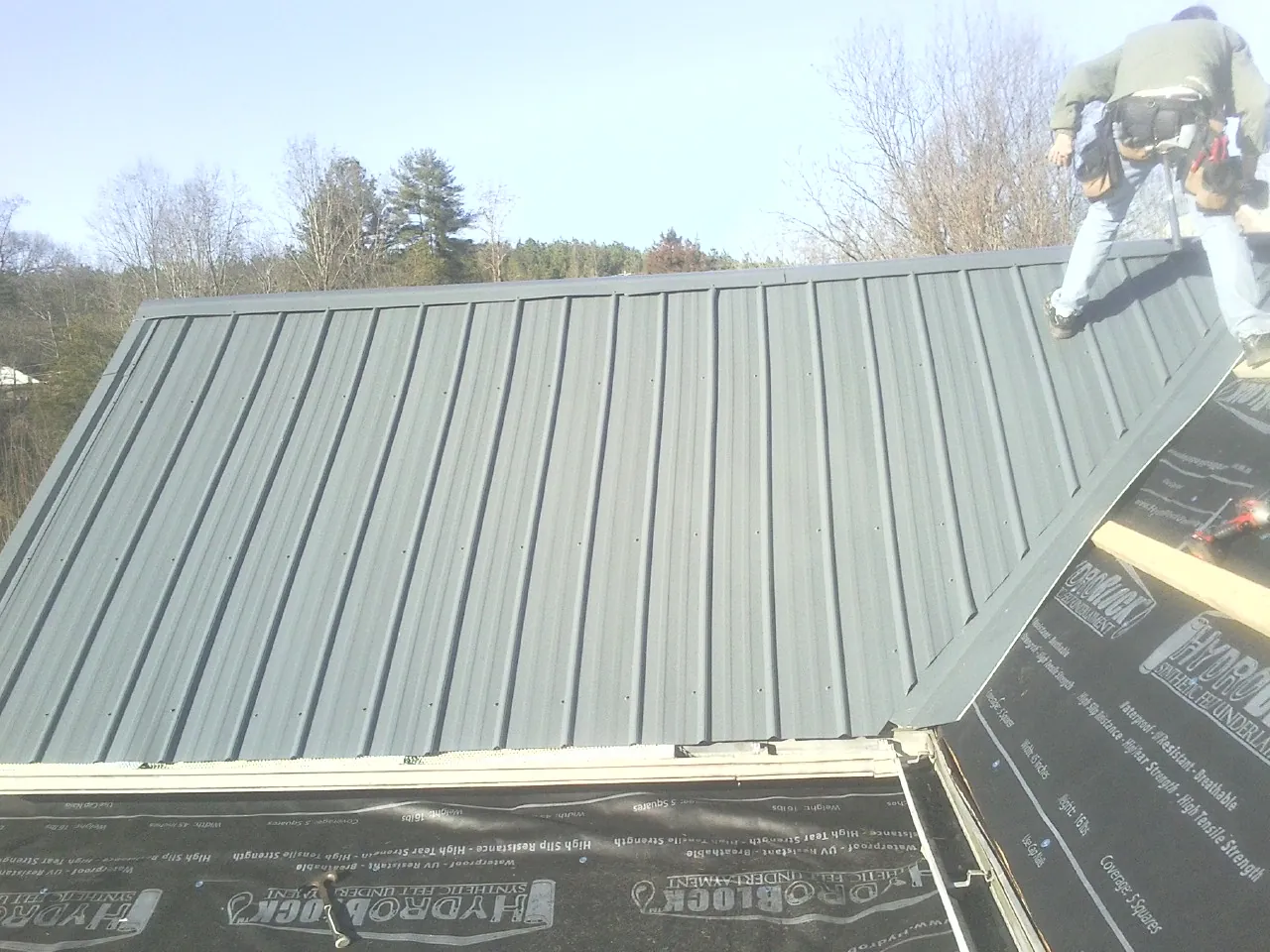 Crew installing dark green standing seam panels over synthetic underlayment in Louisburg